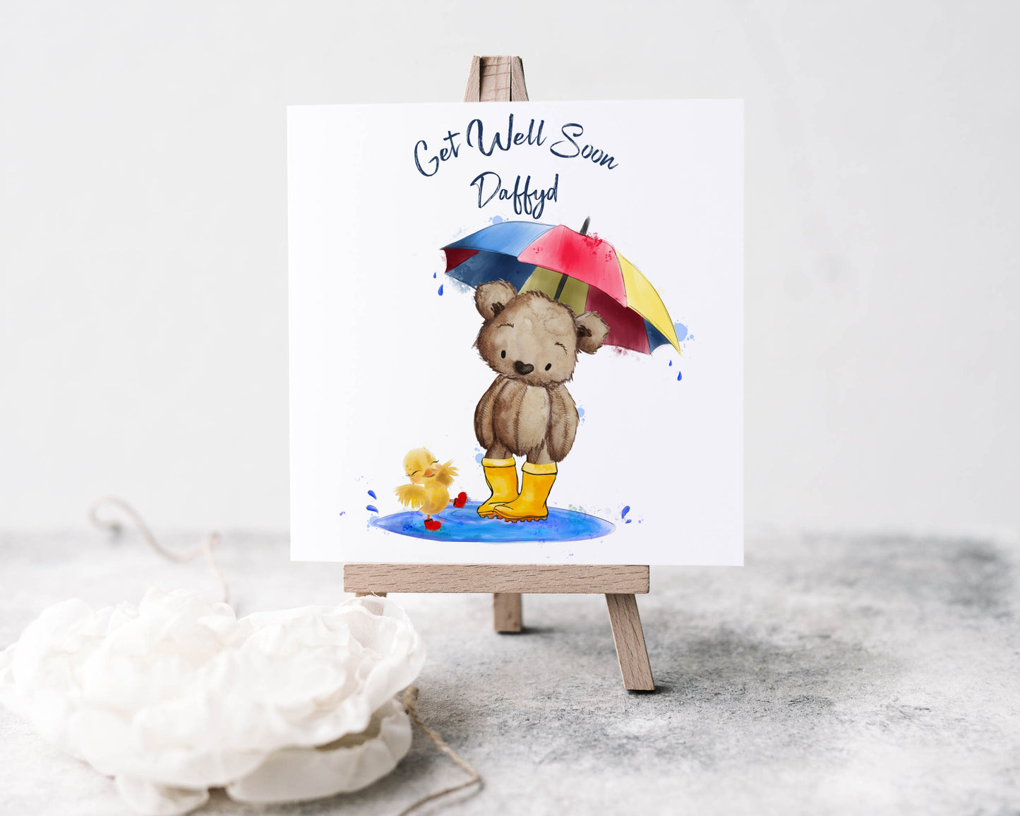 Square card with an image of a teddy bear in wellies holding an umbrella with a chick splashing in a puddle. Text reads 'Get well soon' and is personalised with a name