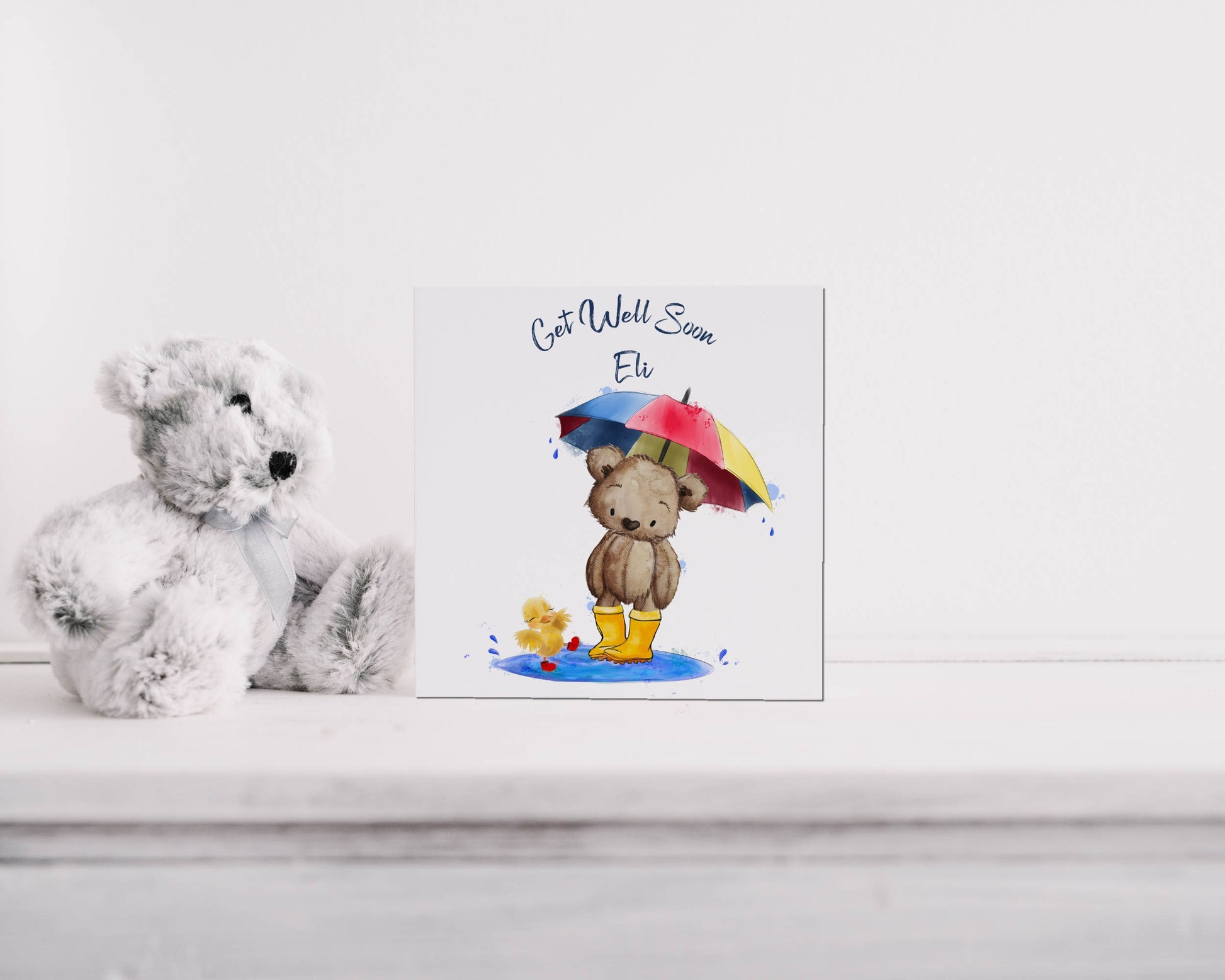 Square card with an image of a teddy bear in wellies holding an umbrella with a chick splashing in a puddle. Text reads 'Get well soon' and is personalised with a name
