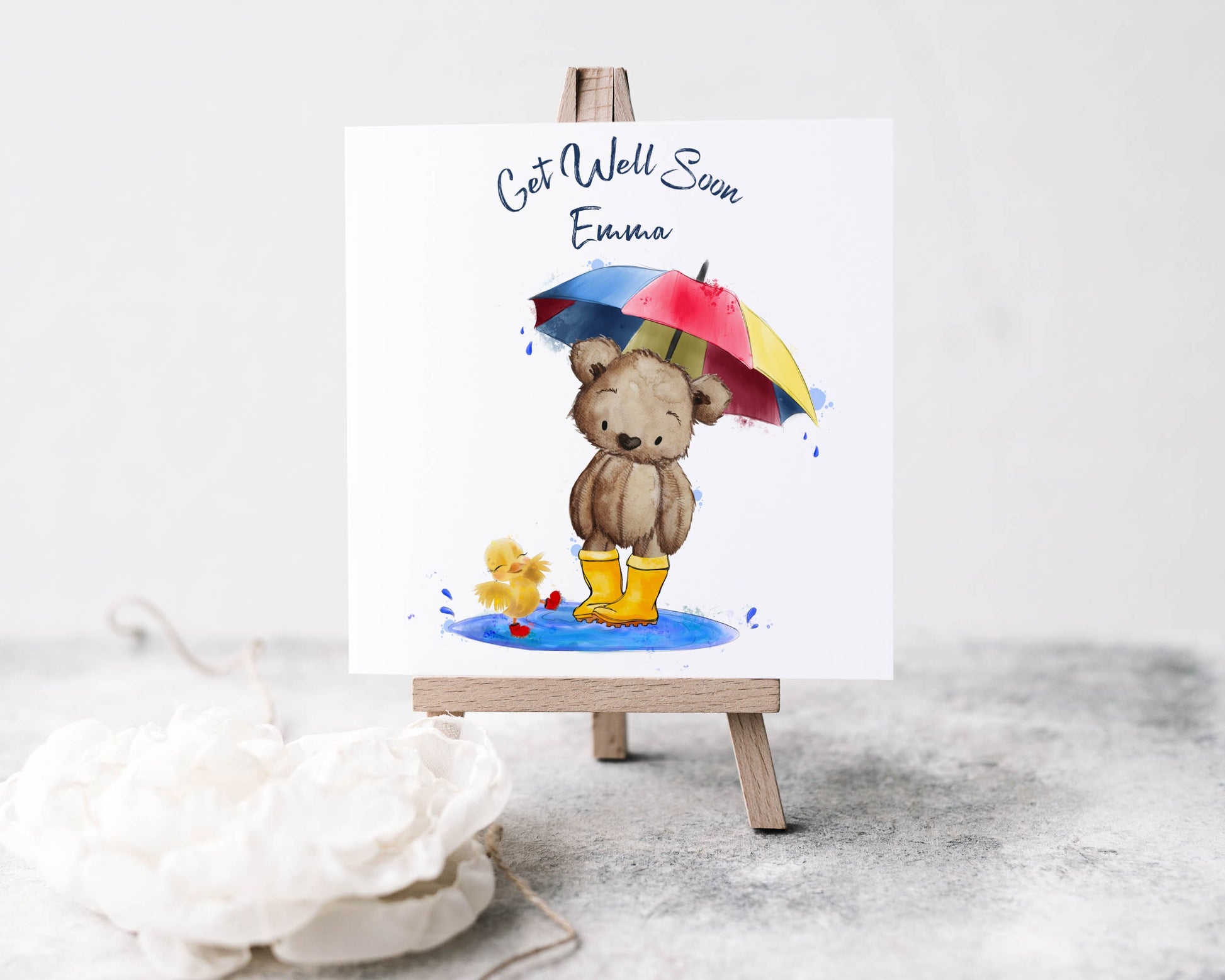 Square card with an image of a teddy bear in wellies holding an umbrella with a chick splashing in a puddle. Text reads 'Get well soon' and is personalised with a name